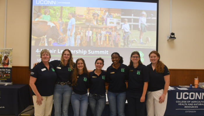 Group of 7 standing and wearing blue UConn 4-H polos behind a powerpoint