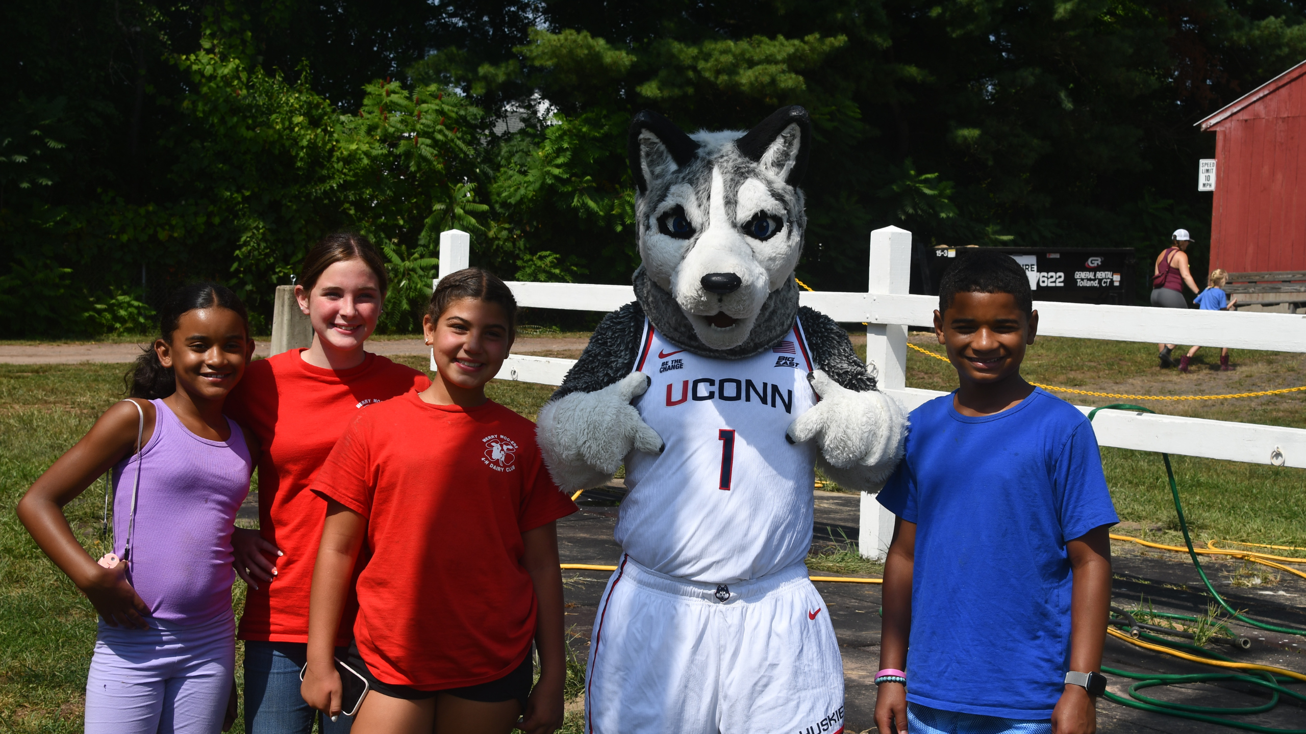 Five youth pose and smile with the UConn Husky mascot outdoors on a sunny day. The mascot is wearing a white UConn basketball jersey with the number 1. The children are casually dressed in colorful summer clothes, standing in front of a white wooden fence with trees and a red barn in the background. The atmosphere is cheerful and festive.