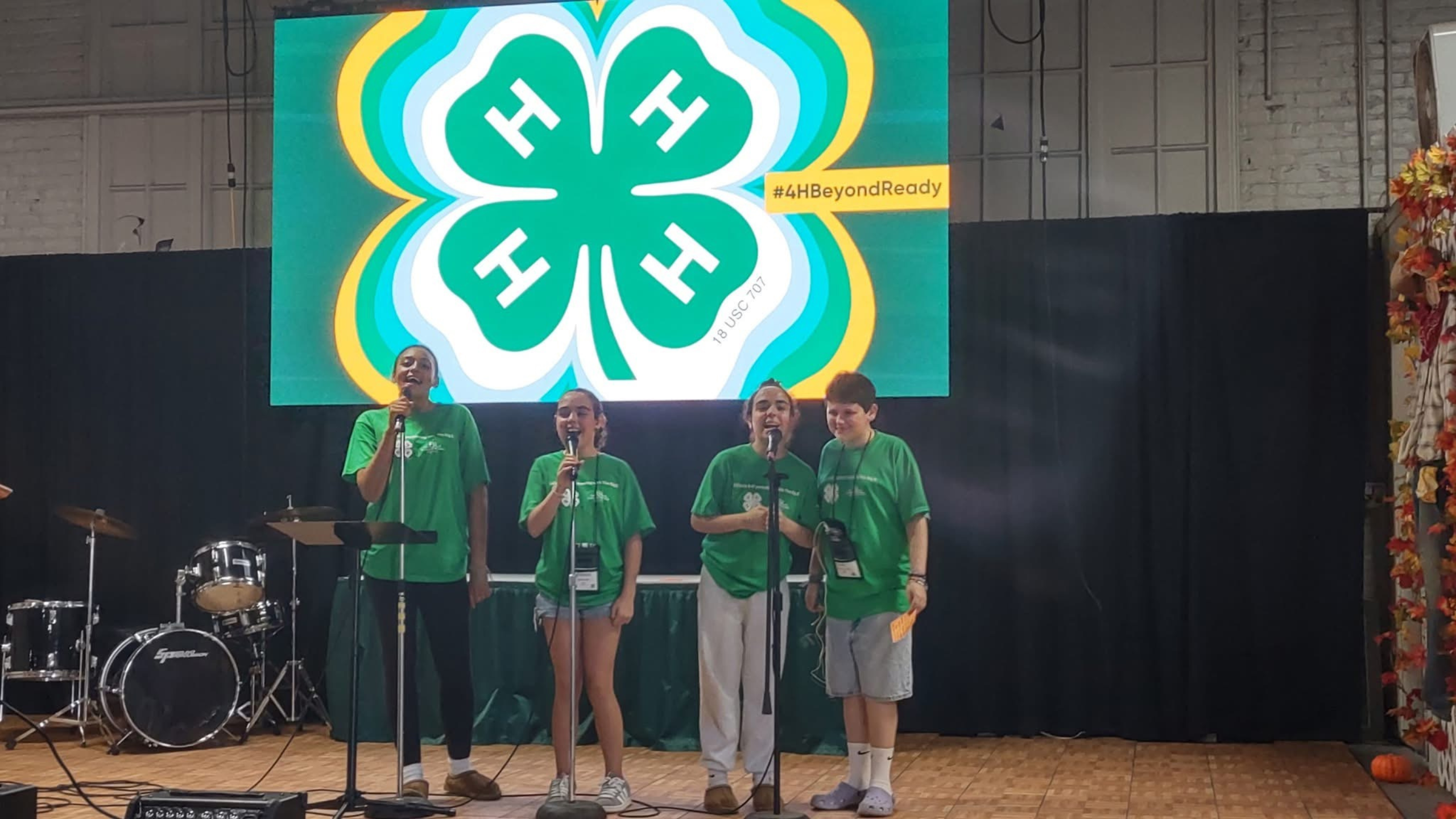 Students standing on a stage with microphones and a 4-H clover in the background