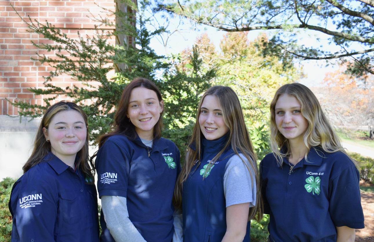 Image of four girls standing in front of a building and trees in blue shirts with a green 4-H clover